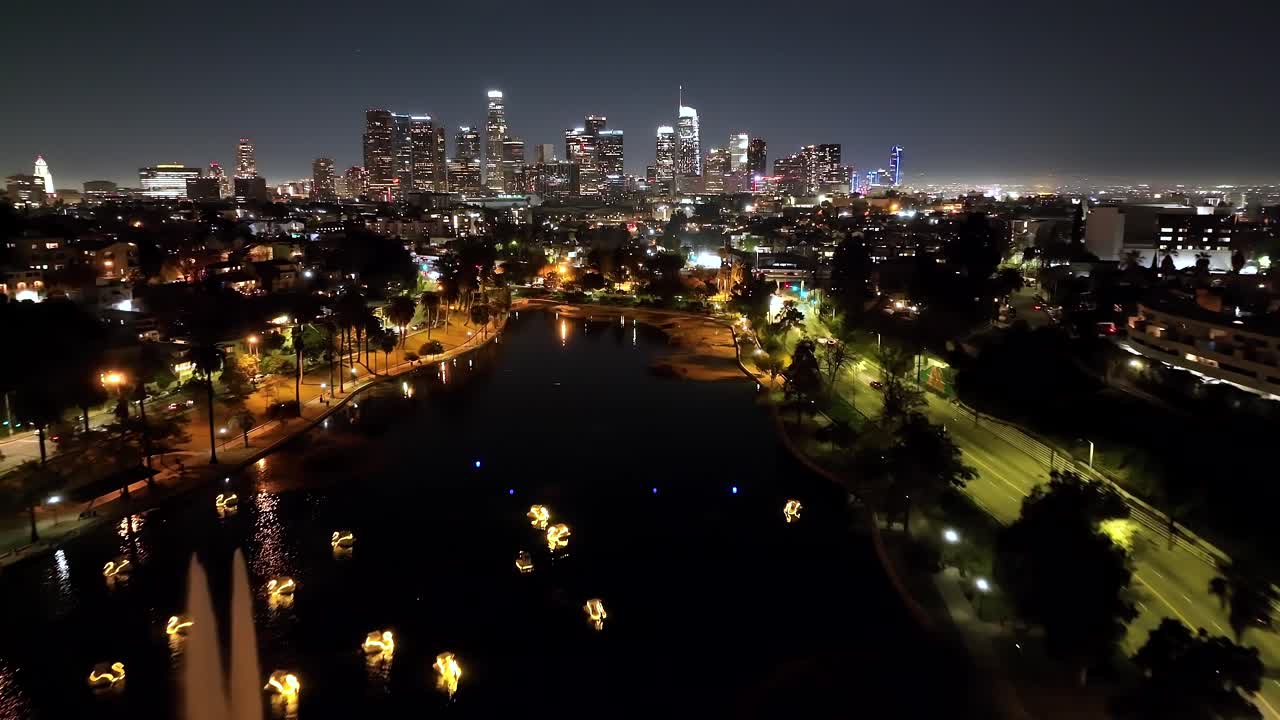 Aerial view flying over Los Angeles Echo park illuminated lake fountain towards futuristic city skyline