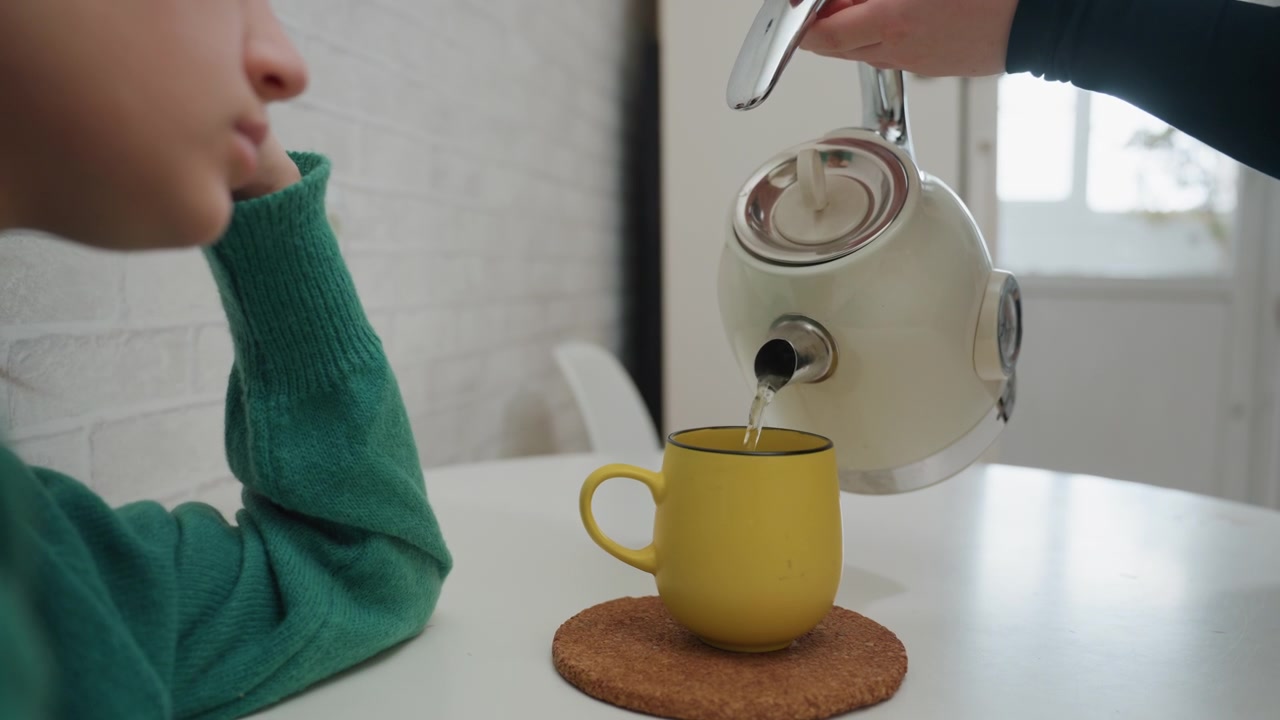 Giovane donna bianca versa il tè in un bollitore nella piccola cucina del nuovo appartamento, versando acqua calda in una tazza gialla mentre appoggia il mento sulla mano, apparendo stanca ma confortata, una pausa nel giorno del trasloco, scena domestica accogliente e calda.