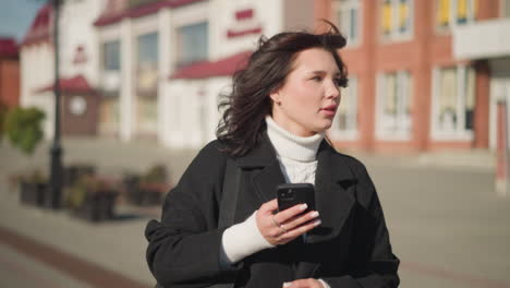 lady walking on urban street holding phone in hand, looking around briefly before focusing back on phone, background features blurred buildings with red and white accents under bright sunlight