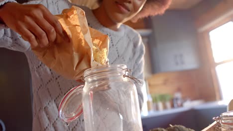 Happy-african-american-woman-pouring-pasta-into-jar-in-sunny-kitchen,-in-slow-motion