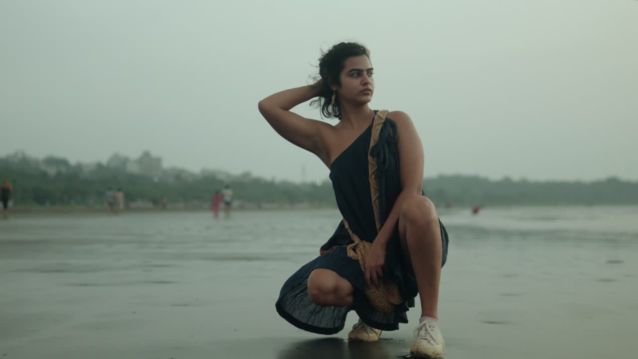 Woman in a black dress crouching on a cloudy beach, reflective mood, with soft focus on distant figures