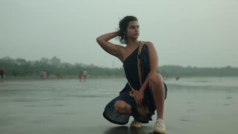 Woman-in-a-black-dress-crouching-on-a-cloudy-beach,-reflective-mood,-with-soft-focus-on-distant-figures