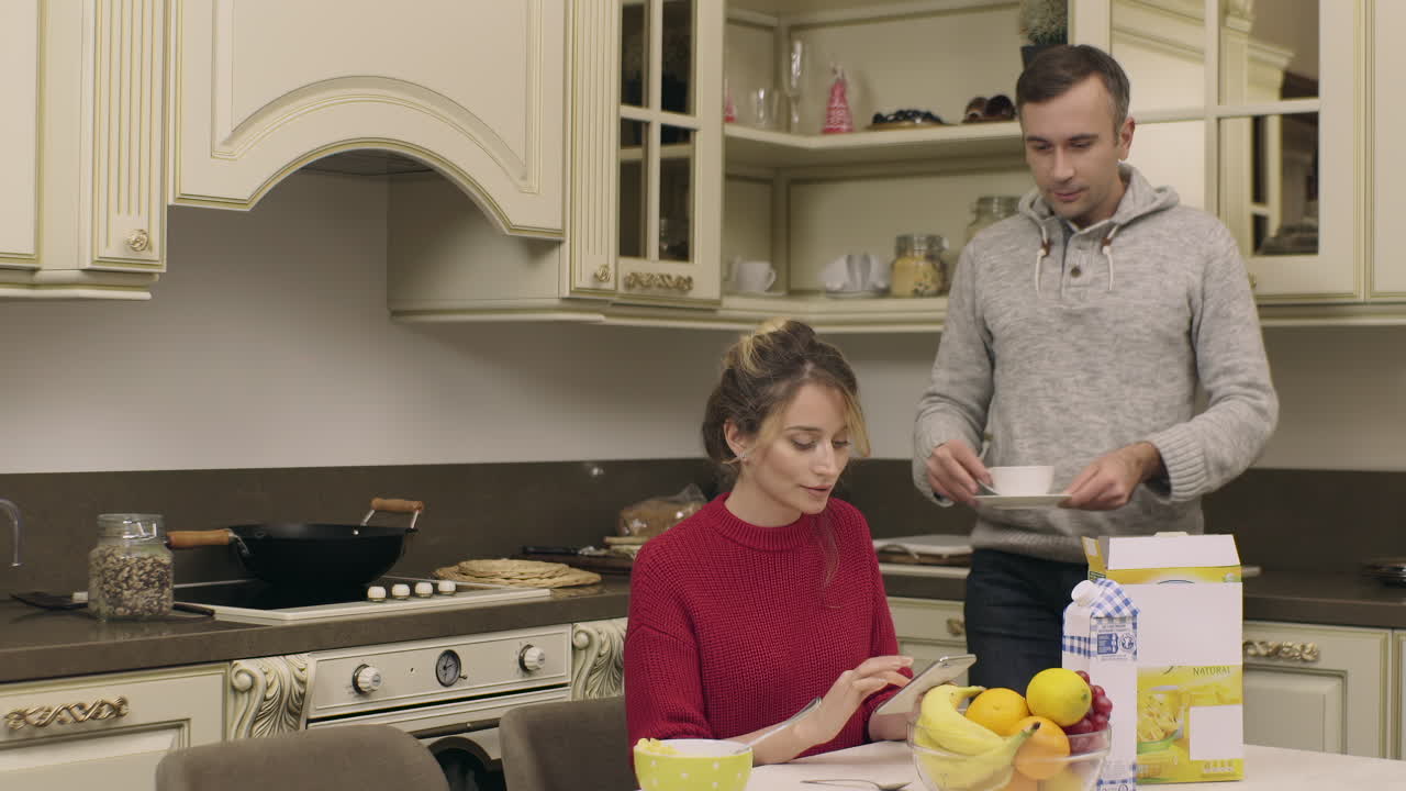 A man serves breakfast to a woman in a modern kitchen. They share a moment together while enjoying their meals. The atmosphere is warm and inviting, filled with delicious food