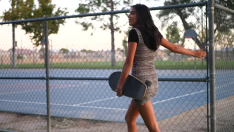 Una-Mujer-Hispana-Muy-Joven-Sonriendo-Y-Caminando-Por-Un-Parque-Urbano-Con-Su-Patineta-A-Lo-Largo-De-Una-Cerca-De-Eslabones-De-Cadena-A-Cámara-Lenta