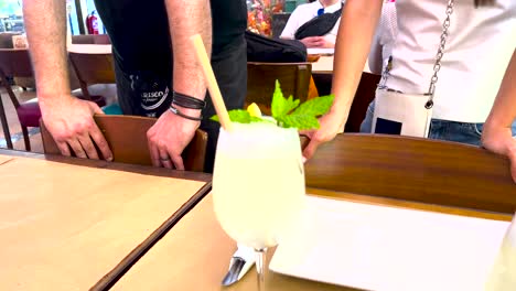 refreshing lemonade with mineral water, zesty lemon slices, fresh mint, and ice, served on a rustic wooden table with people in the background