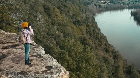 aerial photo of a woman on the edge of a cliff taking landscape photos by a river, she is wearing an orange cap, long traveling forward