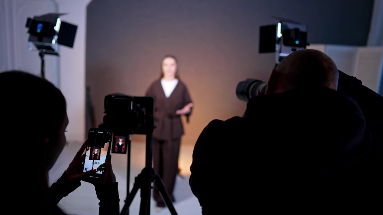 Asistentka natáčí ženu chytrým telefonem během video produkce. Osoba drží telefon vertikálně, aby nahrála video ženy stojící před profesionálním osvětlením ve studiu.
