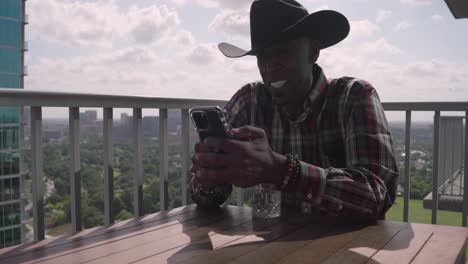 Schwarzer-Mann-Mit-Cowboyhut,-Der-Lächelnd-Und-Lachend-Auf-Das-Telefon-Schaut