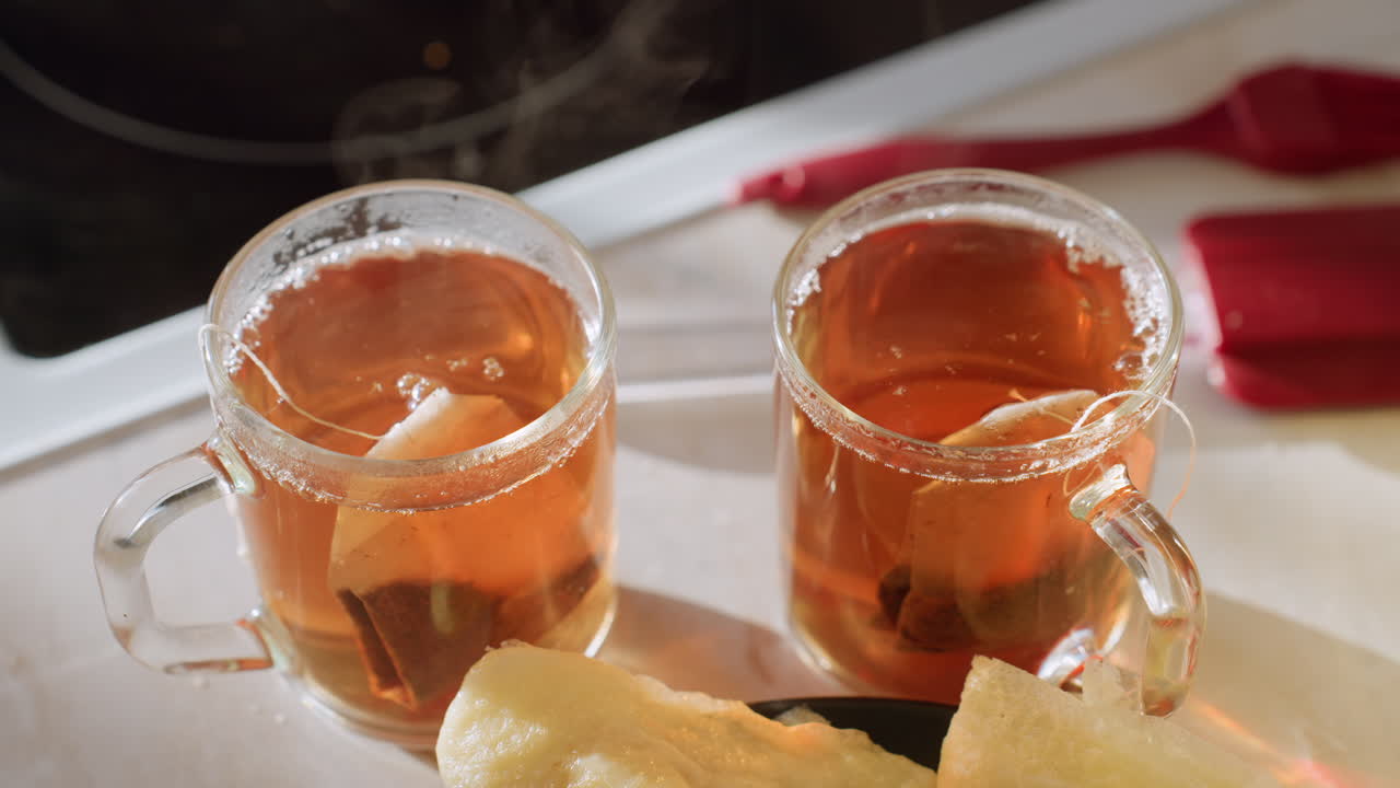 Close up of two steaming glass cups filled with amber tea as teabags steep, placed beside freshly made pancakes on kitchen counter in warm morning light creating cozy breakfast