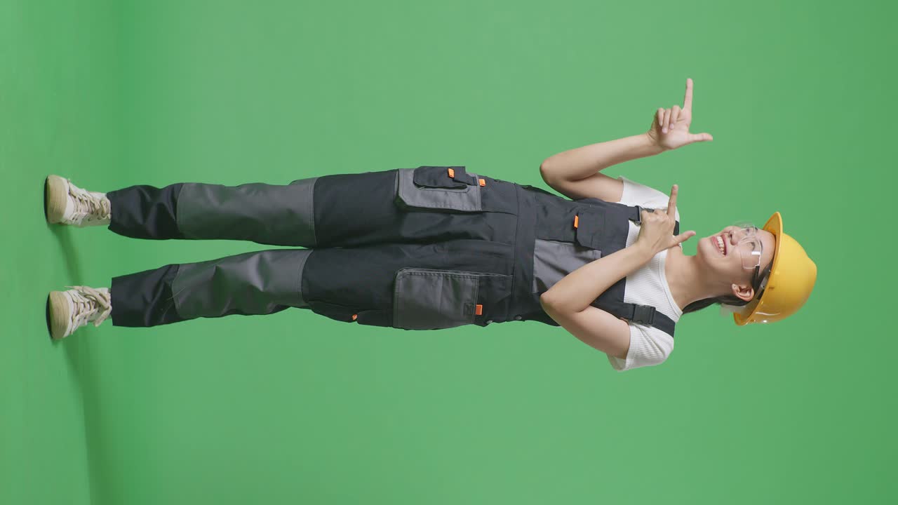 안경과 안전 헬을 입은 아시아 여성 노동자의 몸이 가득 차 있으며, 녹색 스크린 배경 스튜디오에 서서 웃고 을 가리키고 있습니다.