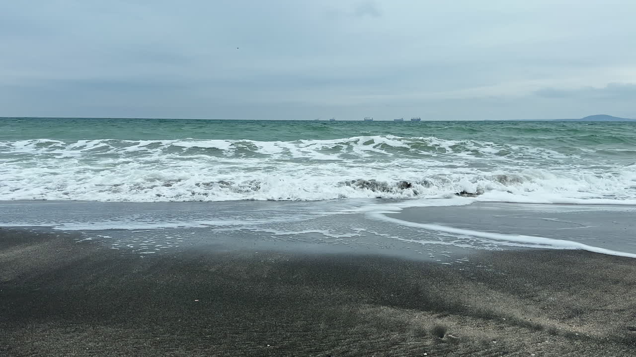 Onde si infrangono su una spiaggia sabbiosa con navi in lontananza durante una giornata nuvolosa lungo la costa