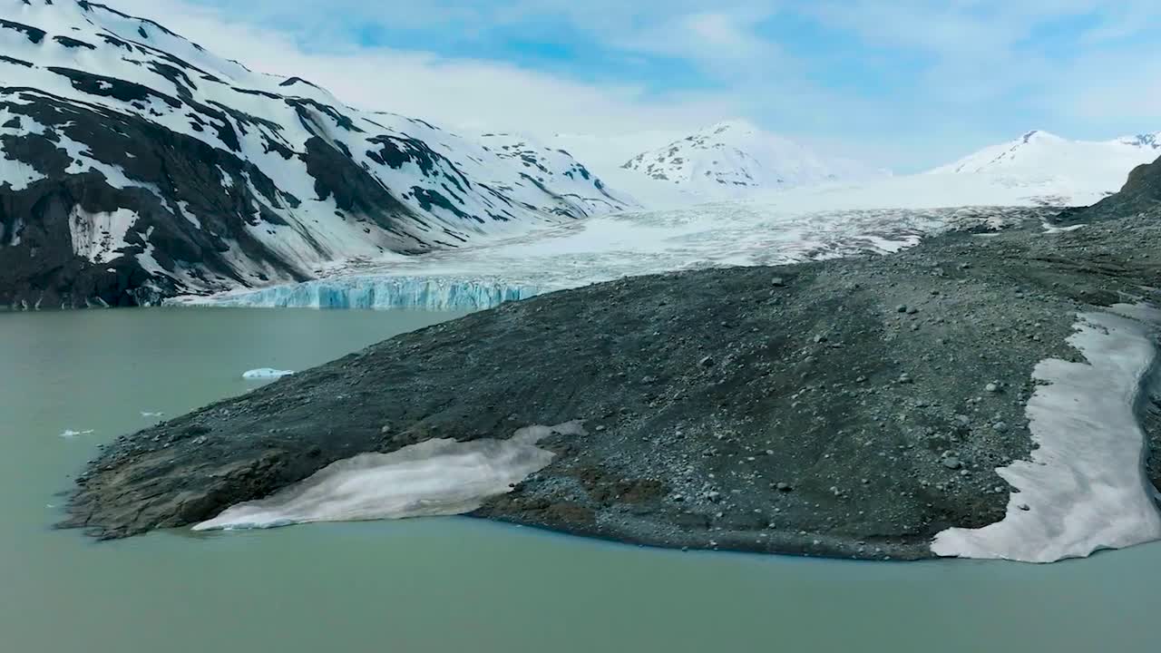Aerial drone gliding with low angle over arctic water around brown and dark black sandy muddy shoreline and a glacier behind it that is between mountains during a sunny winter day. Cold water in front