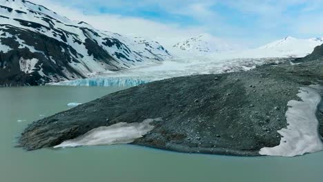 aerial drone gliding with low angle over arctic water around brown and dark black sandy muddy shoreline and a glacier behind it that is between mountains during a sunny winter day. cold water in front