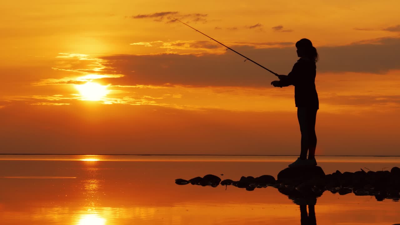Wanita memancing dengan joran pancing berputar dengan latar belakang matahari terbenam.