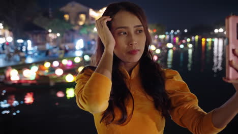 Woman-in-yellow-Ao-Dai-taking-a-selfie-at-Hoi-An's-vibrant-night-scene,-colorful-lights-reflecting-on-water