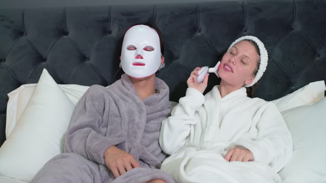 Divers stel in badjassen op bed, man doet LED-masker op, vrouw glijdt met apparaat voor huidverzorging