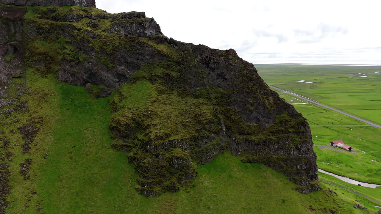 Aerial view of vibrant green fields stretching below steep moss-covered cliffs in Suðurland, Iceland, with winding streams and dramatic shadows shaping the vast volcanic landscape