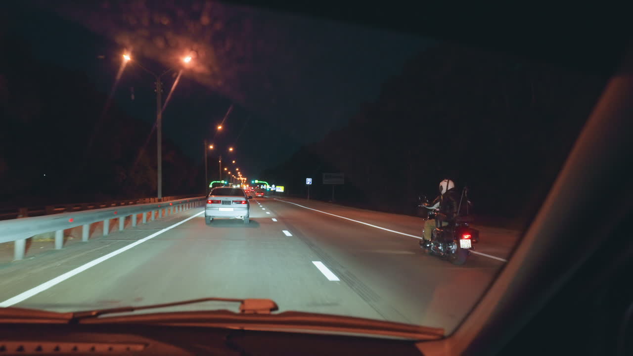 Car and motorcycle move along illuminated night highway viewed from inside another vehicle, showing street lights, road markings, and glowing signs in distance under dark sky