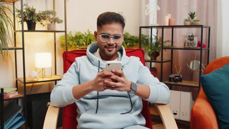 indian arabian young man sitting on chair use smartphone share messages on social media application
