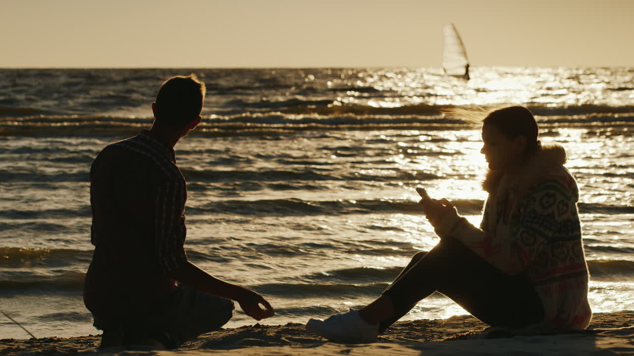 남자가 돌을 던지는 전화를 즐기는 일몰에 해변에서 휴식을 취하는 로맨틱 커플