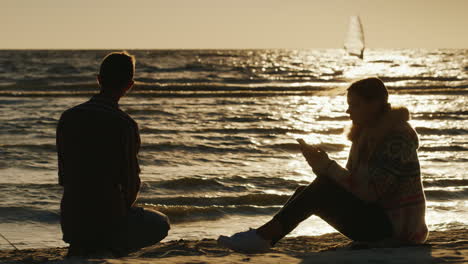 Romantisches-Paar-Entspannt-Am-Strand-Bei-Sonnenuntergang-Frau-Genießt-Ein-Telefon,-In-Das-Ein-Mann-Steine-Wirft