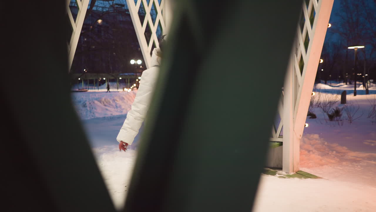 Through lattice of illuminated gazebo, artist wearing white coat and headphones gently moves body within snowy park in soft dusk, framed by wooden lattice, in serene winter dance scene