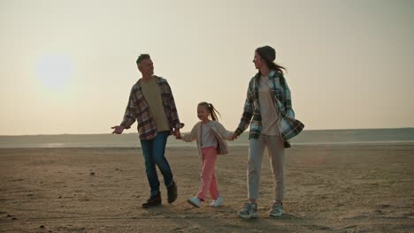 A-happy-brunette-man-in-a-checkered-shirt-walks-hand-in-hand-with-his-little-daughter,-a-blonde-girl-in-a-white-jacket-and-pink-pants,-and-his-wife,-a-brunette-girl-in-a-checkered-green-shirt,-during-his-vacation-outside-the-city-in-the-summer-evening