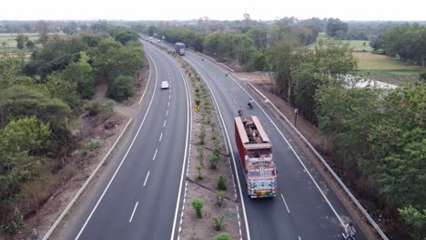 High-angle-shot-of-a-generic-new-well-maintained-national-highway-of-India-with-rural-landscape-of-farmlands-and-lush-green-tree-border-with-trucks-and-passenger-vehicle-movement