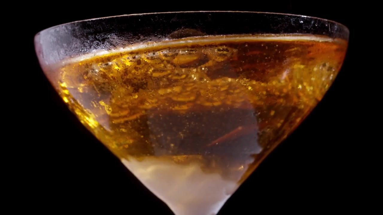 Bicchiere da cocktail ghiacciato che si riempie di liquido ambrato dorato e bollicine che salgono su sfondo nero.