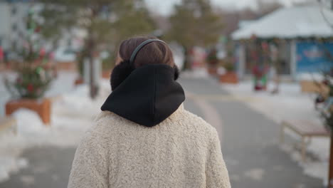 back view of girl wearing warm beige winter coat, black hoodie, and fluffy earmuffs while walking outdoors on snowy day, blurred background with festive decorations, snow-covered ground, and trees