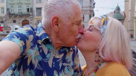 POV-shot-of-senior-tourists-couple-taking-selfie-in-summer-city-center-smiling,-looking-at-camera