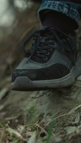 una vista cercana de los pies de una persona con zapatillas negras inclinando una pierna, sentada en una pendiente de hierba, la persona está sosteniendo una pistola negra