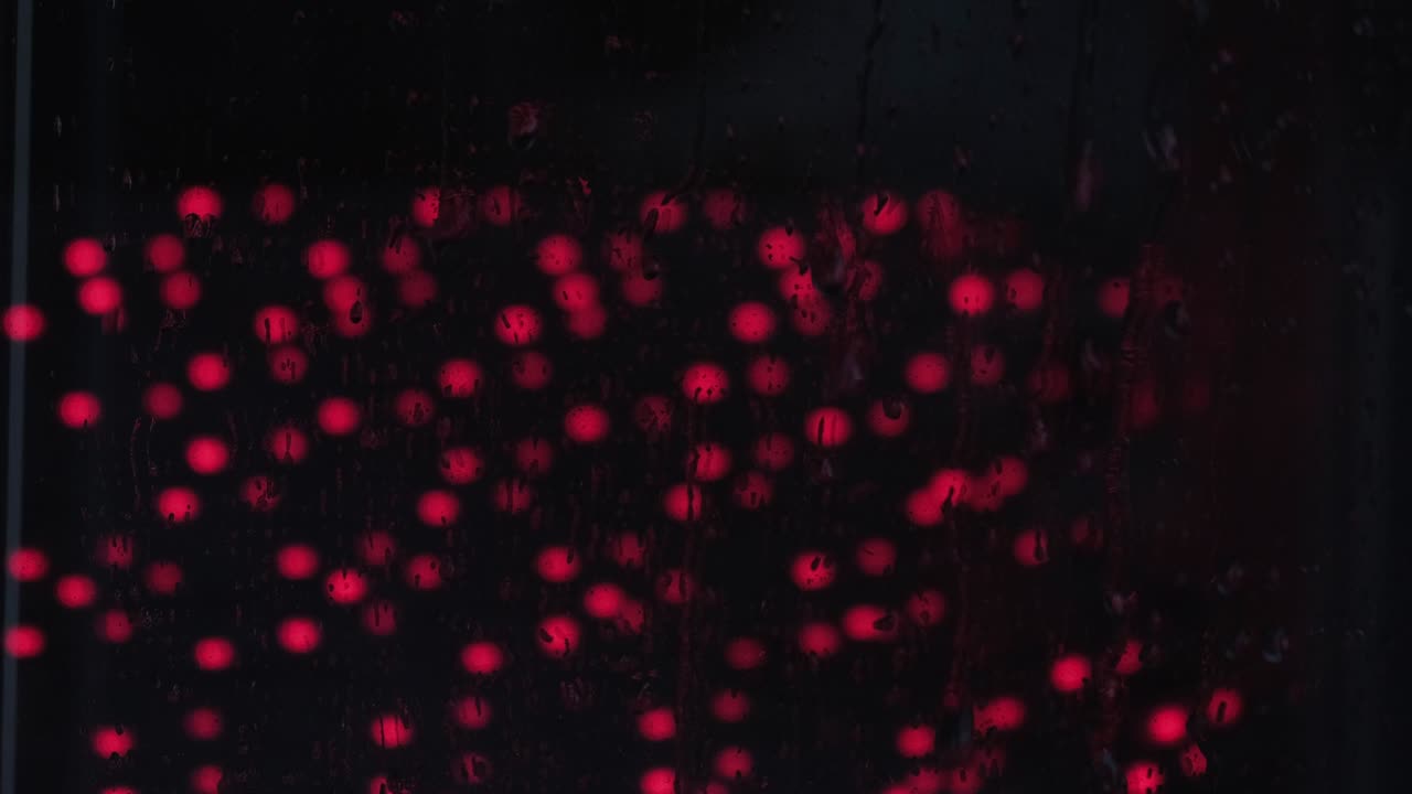 Raindrops on the window glass close-up with Red light. Night light reflection on wet glass. Droplet background texture