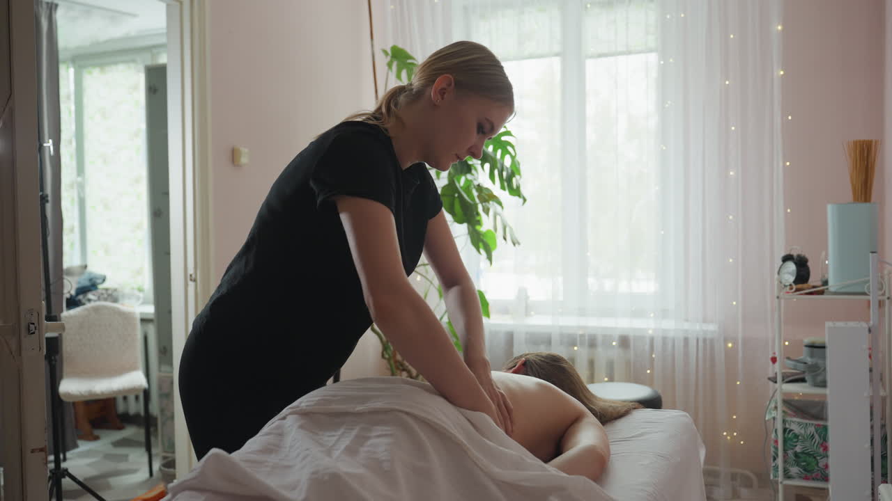 Massage therapist performing rib massage on client in cozy wellness room with indoor plant, sheer curtain, and white chair with black metal leg visible through open doorway in soft natural lighting