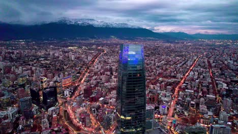 Dolly-In-Der-Luftaufnahme-Des-Costanera-Turms-Und-Des-Geschäftszentrums-Von-Santiago,-Chile-Bei-Nacht-Mit-Den-Schneebedeckten-Und-Bewölkten-Anden-Im-Hintergrund