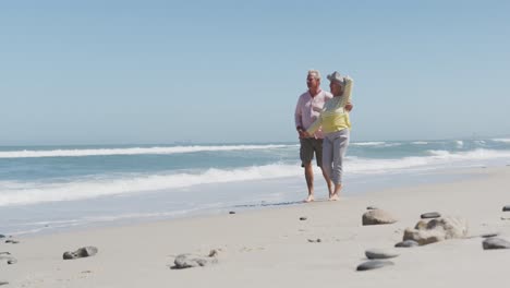feliz pareja caucásica mayor cogida de las manos hablando entre sí mientras caminan por la playa