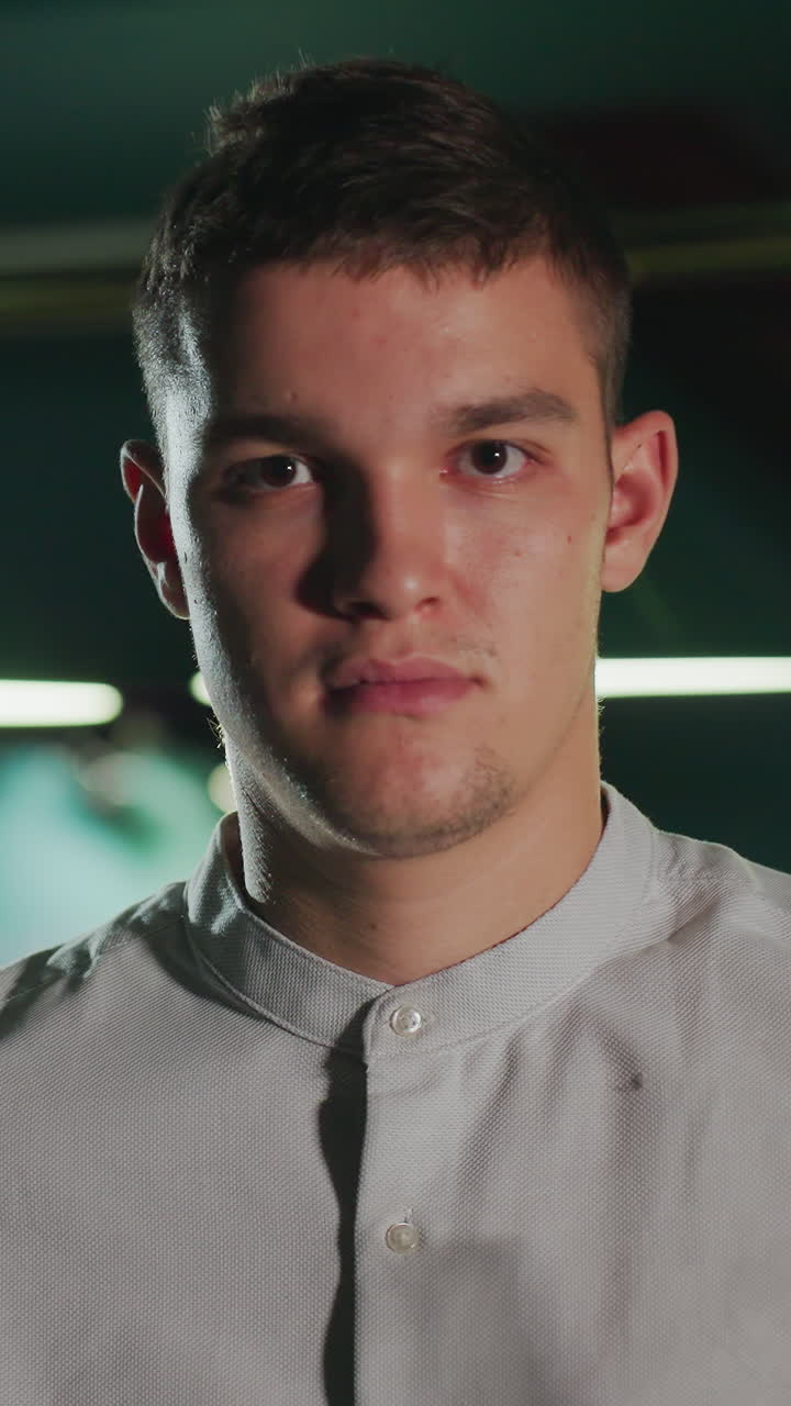 un jugador con camisa blanca se sienta en una posición enfocada, sosteniendo el palo de la señal antes de levantarse con la cara seria en una sala de billar débilmente iluminada.