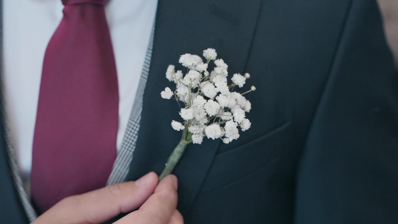 Close up of a hand attaching a small white flower boutonniere to a dark suit jacket