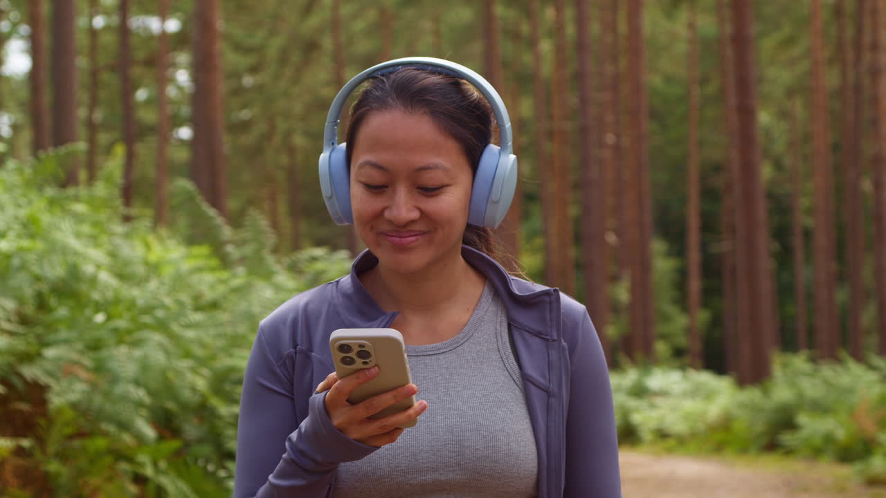 mujer con auriculares inalámbricos haciendo ejercicio haciendo ejercicio al aire libre transmitiendo música o podcast desde un teléfono móvil corriendo por una pista en el bosque con ropa deportiva filmada en tiempo real