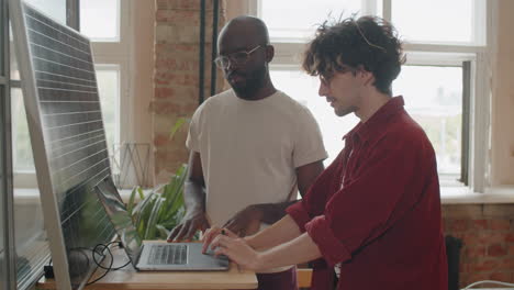 Diverse-Colleagues-Charging-Laptop-with-Solar-Panel-and-Speaking-in-Office