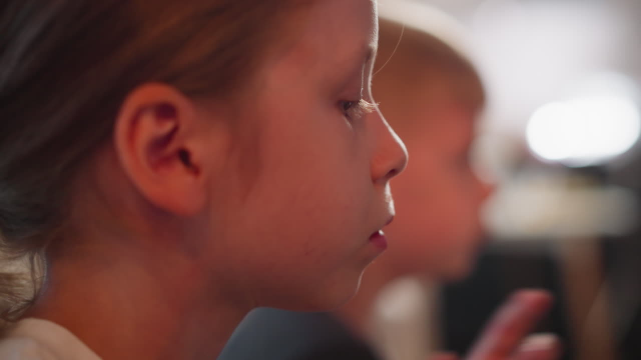 close up of young girl chewing food with focus on facial expression while little boy in background plays around blurred lights and indoor setting during casual moment of eating together at home