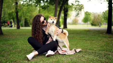 happy girl proud dog owner is caressing and kissing her pet sitting on grass in the park while the animal is enjoying love and care. green trees and lawns are visible.