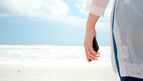 Mid-section-of-woman-standing-with-mobile-phone-at-beach-4k