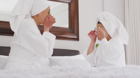 video of happy asian mother and daughter in robes doing cucumber mask and having fun