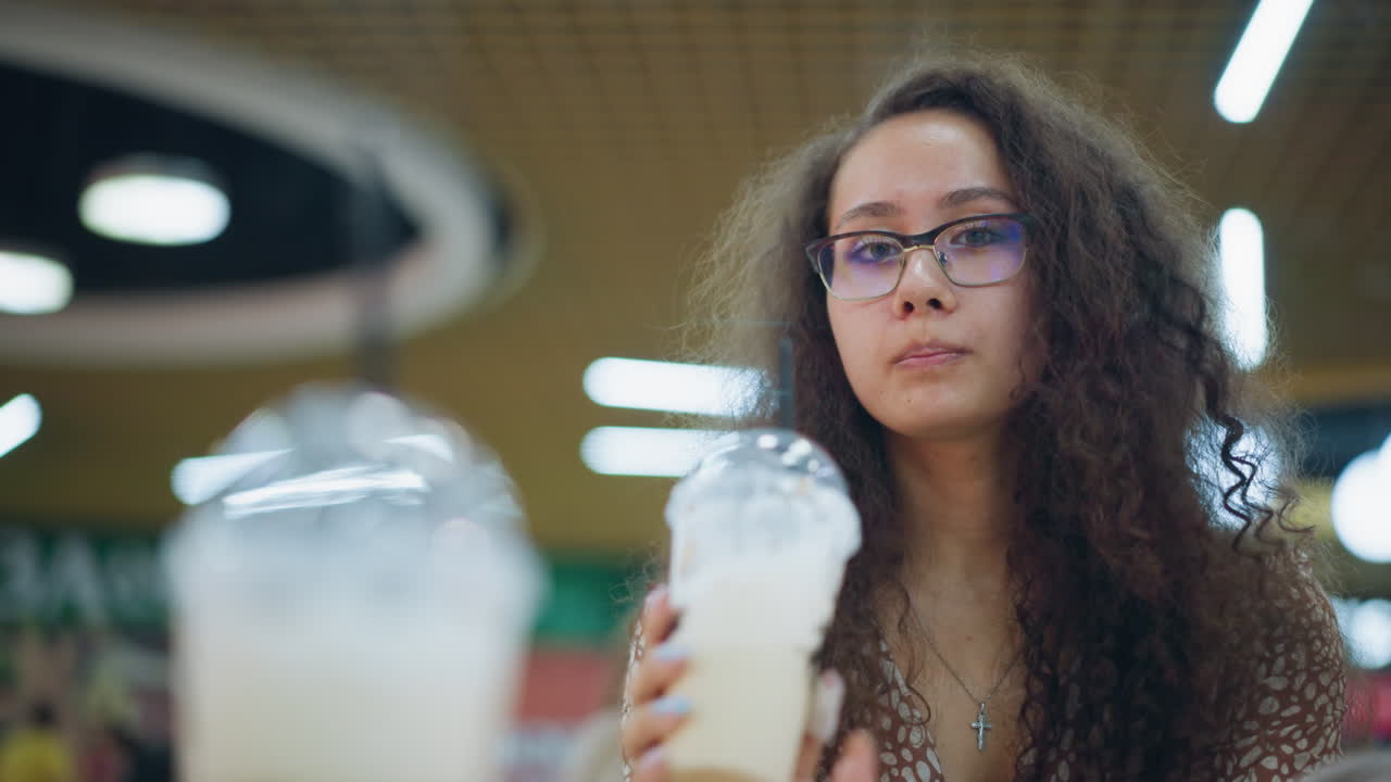 mujer bebiendo una bebida cremosa con paja, sonriendo en un ambiente animado del centro comercial, creando una atmósfera relajada y agradable con fondo borroso, luces bokeh, y otra bebida cerca en la mesa