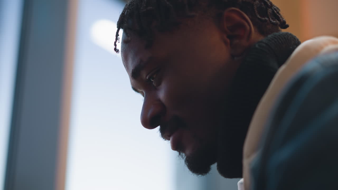 close up side profile of young man with dreadlocks and beard staring down in contemplative mood wearing hoodie under denim jacket illuminated by soft window light in indoor minimal setting