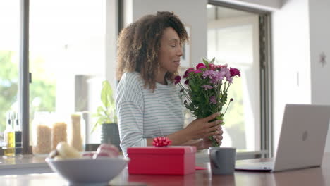 Mujer-Birracial-Feliz-Con-Regalos-Y-Flores-Teniendo-Videollamada-En-Una-Laptop-En-Casa,-Cámara-Lenta