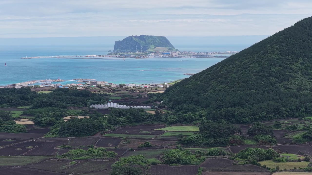 Plano horizontal en cameo con desplazamiento lateral hacia la izquierda a lo largo de Gujwa-eup, Isla de Jeju, Corea del Sur, mostrando tierras de cultivo, pueblo costero, costa volcánica, colinas verdes y una isla volcánica distante
