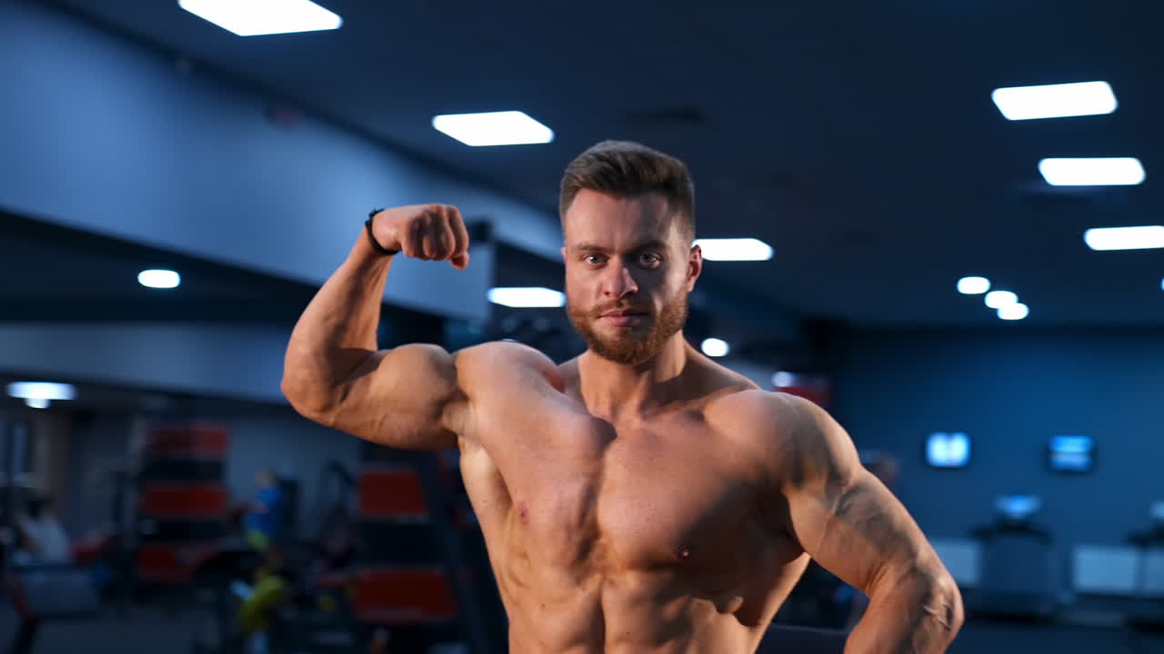 Circle view on bodybuilder without shirt in gym. Man with strong hands and big muscles. Slow motion.