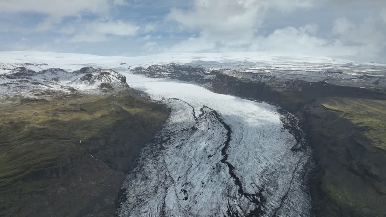 Drone view of the expansive Sólheimajökull glacier in Iceland, flowing between rugged mountains and lush green terrain under a partly cloudy sky.